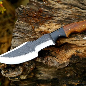 Puede incluir: Un cuchillo de caza hecho a mano con un mango de madera marr&oacute;n oscuro y una virola de metal negro. La hoja est&aacute; hecha de acero y tiene un acabado texturizado.