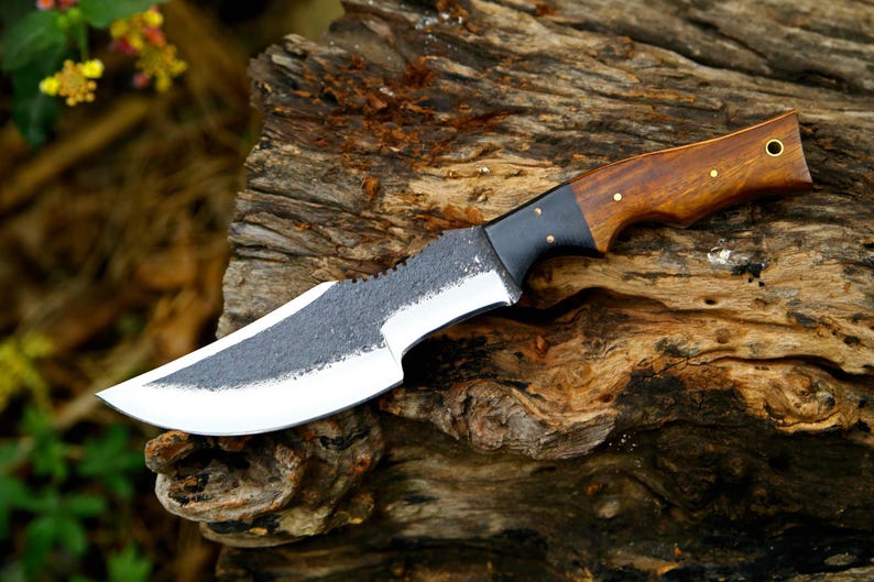 Puede incluir: Un cuchillo de caza hecho a mano con un mango de madera marr&oacute;n oscuro y una virola de metal negro. La hoja est&aacute; hecha de acero y tiene un acabado texturizado.