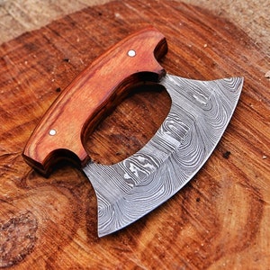 Op de afbeelding: Een ulu-mes van Damascusstaal met een gebogen lemmet en een houten handvat. Het handvat is gemaakt van roodbruin hout en is vastgezet met twee zilveren klinknagels. Het lemmet heeft een uniek patroon en is ontworpen om te hakken en te snijden.