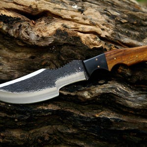 Puede incluir: Un cuchillo con mango negro, mango de madera marr&oacute;n y hoja dentada. El cuchillo est&aacute; apoyado en un trozo de madera desgastada.