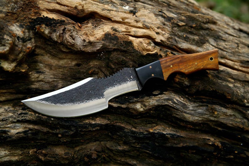 Puede incluir: Un cuchillo con mango negro, mango de madera marr&oacute;n y hoja dentada. El cuchillo est&aacute; apoyado en un trozo de madera desgastada.