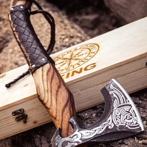 May include: A handcrafted axe with a wooden handle and a dark, patterned leather grip. The axe head features intricate engraved designs. The axe rests on a wooden surface next to a wooden box with the word "Viking" and a compass design.