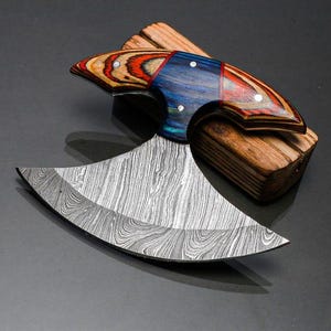 May include: A Damascus steel cleaver knife with a curved blade and a colorful wooden handle. The handle features red, blue, and natural wood tones. The knife rests on a small wooden block, showcasing its craftsmanship.