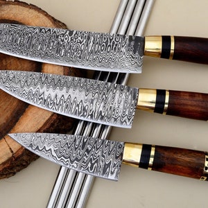 Può includere: Set di tre coltelli da cucina con lame a motivi e manici in legno con accenti dorati. I coltelli sono disposti in fila su una superficie in legno.