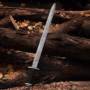 May include: A silver sword with a black leather handle and a decorative pattern on the blade. The sword is lying on a pile of brown logs.