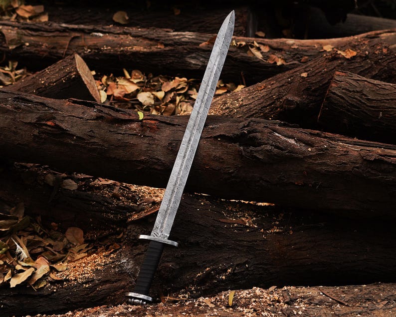 May include: A silver sword with a black leather handle and a decorative pattern on the blade. The sword is lying on a pile of brown logs.
