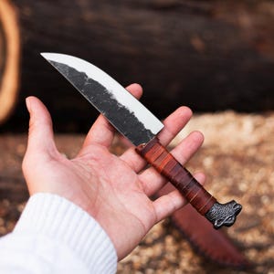 May include: A hand-forged knife with a curved blade and a leather-wrapped handle. The blade has a dark, textured finish with a polished edge. The handle is wrapped in brown leather. The knife is approximately 9 inches long.