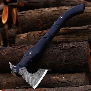 Könnte beinhalten: Eine schwarze Axt mit einem Ledergriff und einer silbernen Klinge. Die Axt liegt auf einem Stapel Holzscheite.