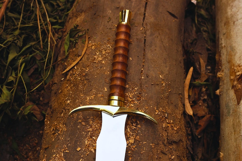 Pu&ograve; includere: Una spada con un manico in legno e una guardia dorata. La lama &egrave; argentata e ha una punta affilata. La spada &egrave; appoggiata su una superficie di legno.