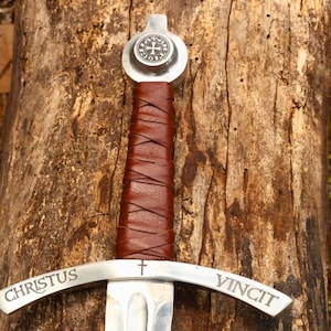 May include: A medieval-style sword with a silver blade and a brown leather-wrapped handle. The crossguard is inscribed with "CHRISTUS VINCIT". The pommel features a circular emblem. The sword rests on a weathered wooden surface.