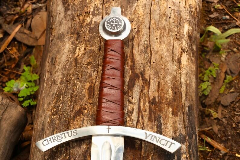 May include: A medieval-style sword with a silver blade and a brown leather-wrapped handle. The crossguard is inscribed with "CHRISTUS VINCIT". The pommel features a circular emblem. The sword rests on a weathered wooden surface.