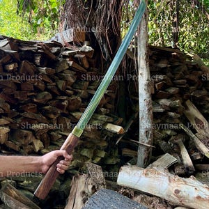 Puede incluir: Una espada larga de color plateado con un mango de madera. La espada está sostenida por una mano frente a una pila de leña apilada.