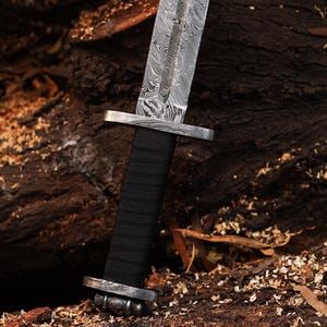 May include: A close-up of a silver sword with a black handle and a patterned blade. The sword is resting on a wooden surface.