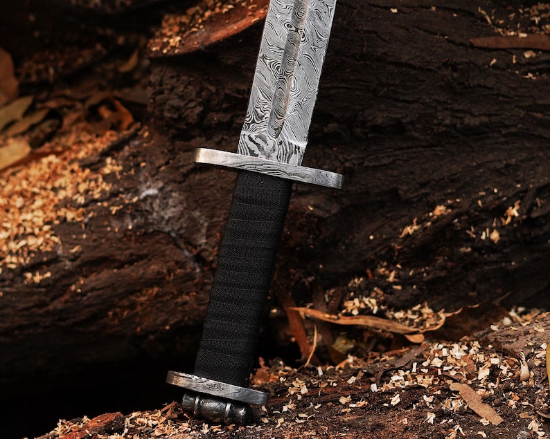 May include: A close-up of a silver sword with a black handle and a patterned blade. The sword is resting on a wooden surface.