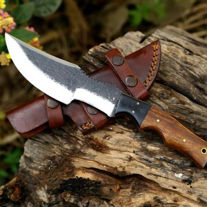 Puede incluir: Una funda de cuero marr&oacute;n con un cuchillo de mango de madera marr&oacute;n y hoja de metal negro. El cuchillo est&aacute; sobre una superficie de madera.