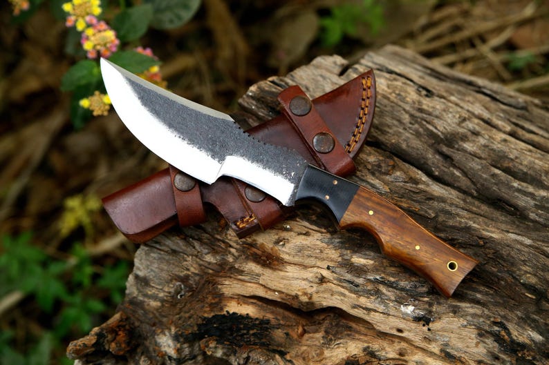 Puede incluir: Una funda de cuero marr&oacute;n con un cuchillo de mango de madera marr&oacute;n y hoja de metal negro. El cuchillo est&aacute; sobre una superficie de madera.