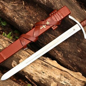 May include: A medieval sword with a silver blade and a brown leather-wrapped handle. The sword rests on a wooden surface next to a brown leather scabbard adorned with gold crosses. The scabbard has straps and a belt loop.