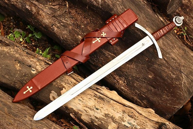 May include: A medieval sword with a silver blade and a brown leather-wrapped handle. The sword rests on a wooden surface next to a brown leather scabbard adorned with gold crosses. The scabbard has straps and a belt loop.