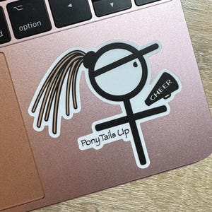 May include: Black and white sticker of a stick figure girl with a ponytail, wearing a hat and holding a megaphone with the word "CHEER" on it. The text "PonyTails Up" is below the figure.