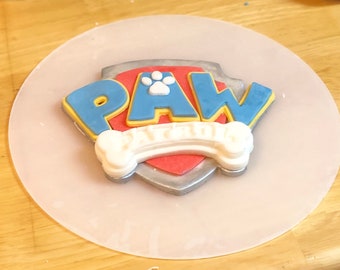 Könnte beinhalten: Ein weißes, rotes, blaues und gelbes Paw Patrol Logo mit den Worten "PAW PATROL" auf einer weißen Kuchen-Dekorationsfläche.