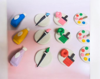 May include: A collection of art-themed decorations in various colours. The image shows miniature paint tubes in blue, yellow, and lavender, alongside paintbrushes, palettes, and other art supplies. The items are arranged on a white and pink background.