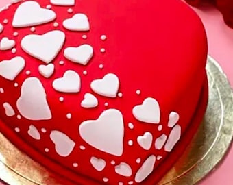 Puede incluir: Un pastel en forma de corazón con glaseado rojo brillante, adornado con decoraciones blancas en forma de corazón de varios tamaños. El pastel está sobre una base redonda dorada, perfecto para una ocasión romántica.