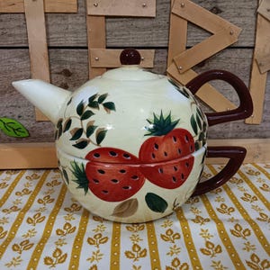 May include: Cream-colored ceramic teapot with a lid and a dark brown handle. The teapot is decorated with large red strawberries and green leaves. The teapot sits on a yellow and white floral patterned tablecloth.