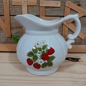May include: White ceramic pitcher with a rounded body and a curved handle. The pitcher is decorated with a strawberry and flower design in red, green, and white. The pitcher has a spout for pouring.