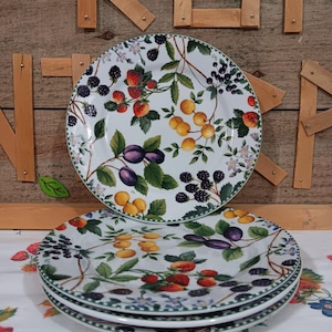 May include: A stack of white ceramic plates with a fruit design. The plates feature illustrations of strawberries, blackberries, plums, and yellow fruit with green leaves. The plates have a green border.