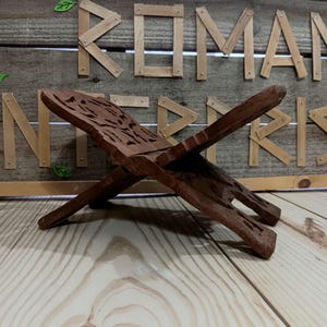 May include: A dark brown wooden book stand with an intricate carved design on the top. The stand is in an open, angled position, and is made of a rich, dark wood. The background features wooden letters spelling out a word.