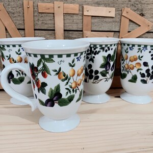 May include: Set of four white ceramic mugs with fruit and leaf designs. Each mug features a different fruit pattern, including plums, strawberries, and yellow berries, with green trim and a white handle. The mugs are on a light wood surface.