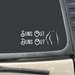 May include: A white vinyl decal with the text "Suns Out Buns Out" on a black car window.