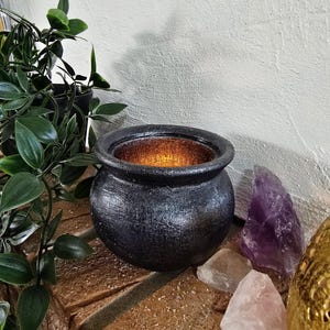 Könnte beinhalten: Ein schwarzer Kessel mit strukturierter Oberfläche, von innen beleuchtet, steht auf einer Ziegeloberfläche. Der Kessel ist von Grün, Kristallen und einem dekorativen goldenen Objekt umgeben und schafft eine mystische und dekorative Präsentation.