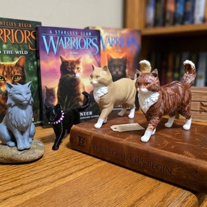 May include: A collection of cat figurines, including a gray, black, tan, and brown tabby cat, displayed with "Warriors" book series. The books and figurines are arranged on a wooden surface, creating a themed display.