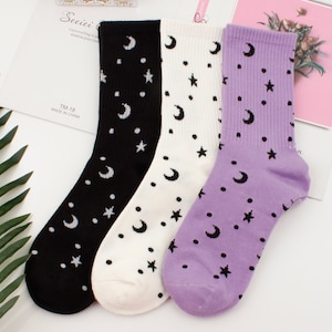 May include: Three pairs of socks with a moon and star pattern. One pair is black with white moons and stars, one pair is white with black moons and stars, and one pair is purple with black moons and stars.