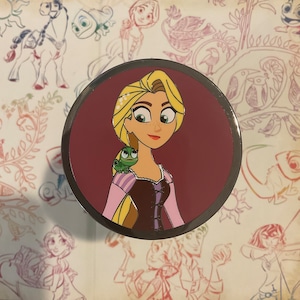 May include: A round pin with a burgundy background featuring a cartoon image of a woman with long blonde hair and a green chameleon on her shoulder. The woman is wearing a purple and brown dress.