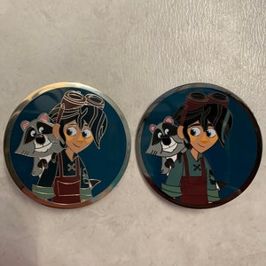 May include: Two round metal pins featuring a cartoon character with a raccoon on their shoulder. The character is wearing a brown vest, blue shirt, and goggles. The background is blue.