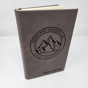 May include: A grey leather-bound journal with a black logo that reads "Resilient Designs LLC, Est. 2021, Custom Laser Engraving". The name "Evan Harrell" is engraved on the front cover.
