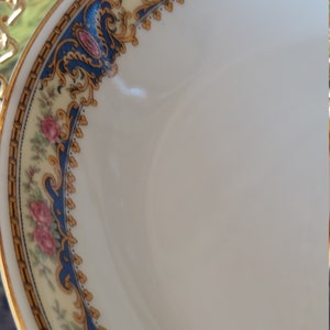 以下が含まれることがあります： 金と青のフローラルな縁取りが施された白い磁器のボウル。縁取りには、ピンクのバラと金のアクセントが施された繊細な花柄が描かれています。