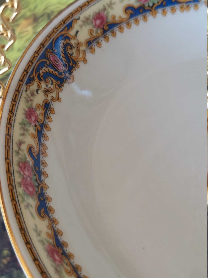 以下が含まれることがあります： 金と青のフローラルな縁取りが施された白い磁器のボウル。縁取りには、ピンクのバラと金のアクセントが施された繊細な花柄が描かれています。