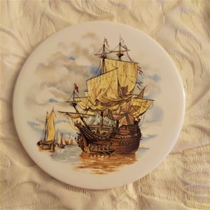 May include: A white ceramic round plate with a painted image of a large sailing ship with brown sails and a brown hull. The ship is sailing on a blue sea with white clouds in the sky.
