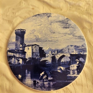 May include: A blue and white ceramic plate with a painted image of a bridge and buildings in a town. The scene is set against a cloudy sky.