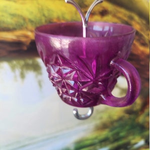 May include: A purple glass teacup with a diamond pattern hangs from a silver hook. The teacup has a handle and is suspended in front of a blurred background.