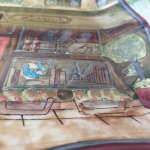 May include: A hand-painted ceramic dish with a whimsical design of a storefront with a red door, bookshelves, a globe, and a potted plant. The dish has a brown and white background with a red border.