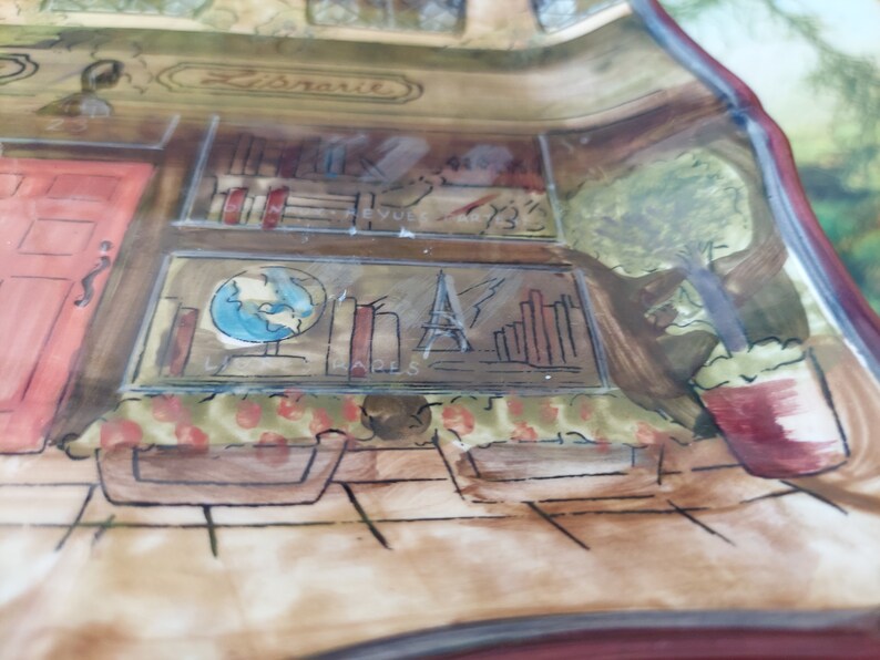 May include: A hand-painted ceramic dish with a whimsical design of a storefront with a red door, bookshelves, a globe, and a potted plant. The dish has a brown and white background with a red border.