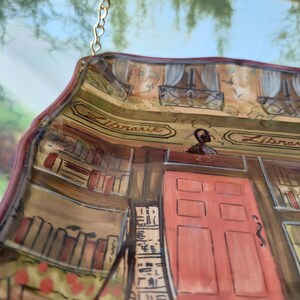 May include: A ceramic wall hanging shaped like a storefront with the word "Librarie" written on the sign. The storefront has a red door with six panels and a window with white curtains. The interior of the store is painted to look like a library with bookshelves filled with books.