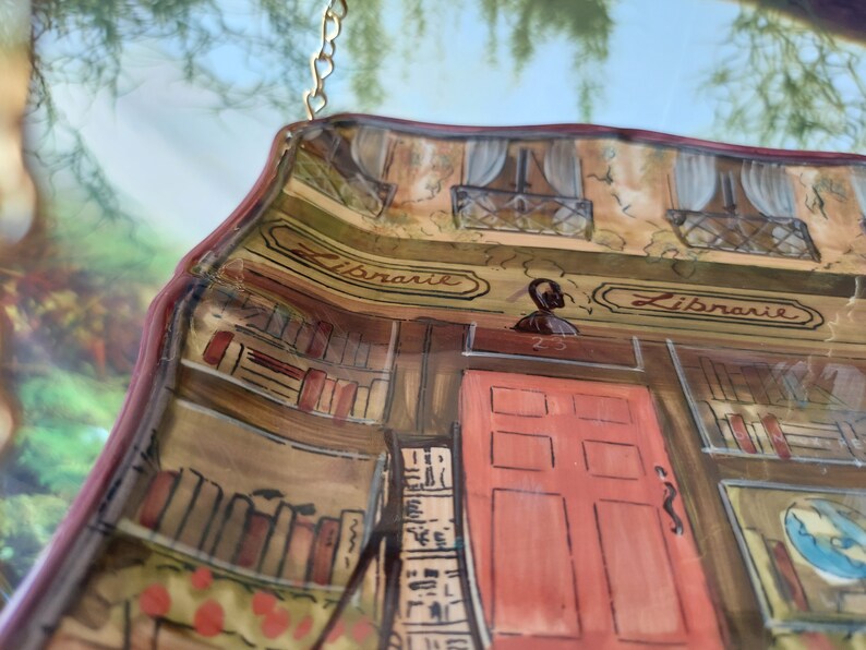 May include: A ceramic wall hanging shaped like a storefront with the word "Librarie" written on the sign. The storefront has a red door with six panels and a window with white curtains. The interior of the store is painted to look like a library with bookshelves filled with books.