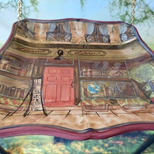 May include: A decorative hanging plate with a painted image of a storefront with the word "Librarie" on the sign. The storefront has a red door and windows filled with books. The plate has a brown and red border.