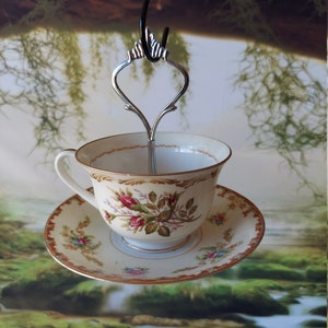 May include: A white teacup and saucer with a floral design hangs from a metal hook. The cup and saucer are decorated with a pattern of pink roses and green leaves on a cream background. The saucer has a gold trim.