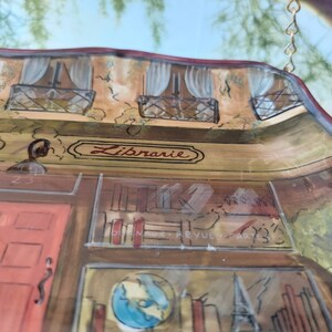 May include: A hand-painted ceramic wall hanging depicting a French bookstore with the word "Librairie" above the door. The bookstore has a red door, bookshelves, and a globe. The hanging is decorated with a gold chain.